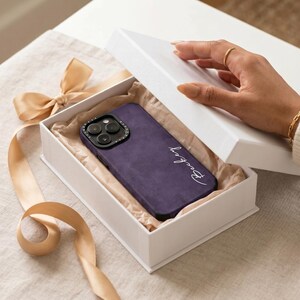 May include: A purple phone case with the name "Bentley" in white script is presented in a white gift box. The case has a textured appearance and a black camera module. A hand is opening the box, revealing the case.