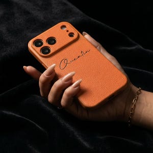 Puede incluir: Funda de teléfono naranja texturizada con el nombre "Quentin" en escritura negra. La funda tiene un recorte para la cámara. Se ve una pulsera de cadena dorada en la muñeca.