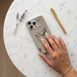 Pode incluir: Uma capa de telefone prateada com o nome "Claire" em escrita preta. Uma caneta e uma pulseira douradas estão ao lado da capa do telefone. Dois raminhos de lavanda estão no lado esquerdo.