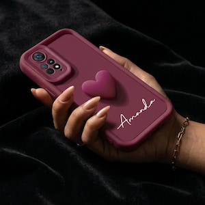 Könnte beinhalten: Eine weinrote Handyhülle mit einem erhabenen Herz und dem Namen "Amanda" in weißer Schrift. Die Hülle hat einen Kameraausschnitt und wird in einer Hand mit einem goldenen Armband gehalten.