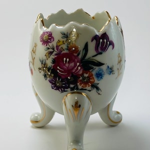 May include: A white porcelain egg-shaped vessel with a scalloped rim and three curved legs. The exterior features hand-painted floral motifs in shades of pink, purple, orange, and blue, accented with gold trim along the rim and the legs.