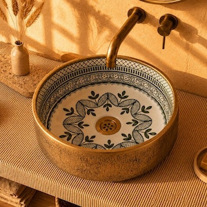 May include: A round ceramic vessel sink featuring a white interior with a dark green botanical pattern and a metallic gold exterior finish. The sink includes a gold drain and is paired with a matching gold wall-mounted faucet fixture.