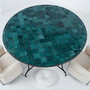 Peut inclure: Une table à manger ronde avec un plateau en carrelage bleu sarcelle foncé à motif d'écailles et une base en métal noir, accompagnée de deux fauteuils rembourrés couleur crème et d'un petit coussin de sol rond blanc capitonné.
