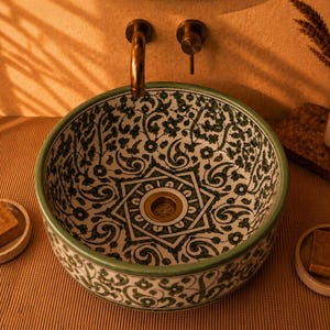 May include: A round ceramic vessel sink featuring a white base with intricate green floral and geometric patterns. The basin includes a central brass drain and is accompanied by two small matching soap dishes, each holding a square bar of soap.