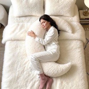 May include: A large, crescent-shaped white faux fur body pillow resting on a matching white faux fur bedspread. The pillow features a soft, textured surface and is positioned on a bed with four white faux fur pillows arranged at the headboard.