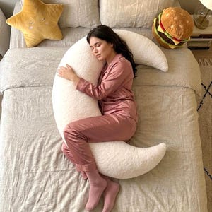 May include: A woman in pink pajamas rests on a bed with a large, white crescent moon-shaped pillow. A yellow star-shaped pillow and a burger-shaped pillow are also on the bed. The bedding is a neutral color.