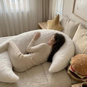 May include: A person in beige loungewear lies on a large, white, fluffy U-shaped body pillow on a bed. A star-shaped pillow and a plush hamburger pillow are also visible. The room has neutral tones and soft lighting.