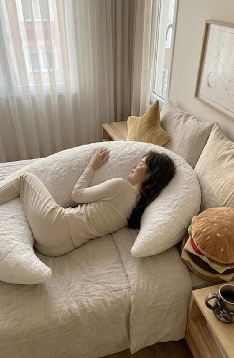 May include: A person in beige loungewear lies on a large, white, fluffy U-shaped body pillow on a bed. A star-shaped pillow and a plush hamburger pillow are also visible. The room has neutral tones and soft lighting.