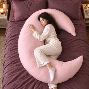 Puede incluir: Una almohada corporal grande en forma de media luna de color rosa claro sólido. La almohada está diseñada para curvarse alrededor del cuerpo, brindando soporte para la cabeza, la espalda y las piernas sobre una cama con ropa de cama morada.