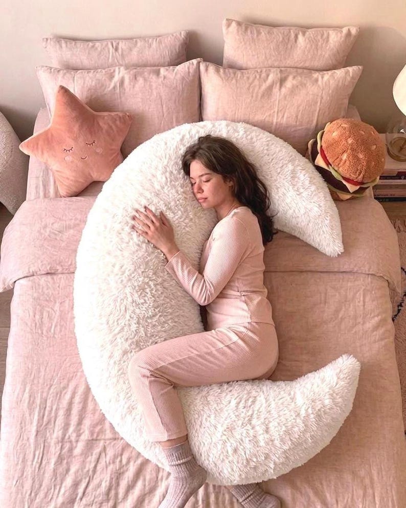 May include: A person is sleeping on a bed with pink bedding, a white crescent moon-shaped pillow, and a star-shaped pillow. A plush burger pillow is also on the bed. The person is wearing a pink long-sleeved shirt and pants.