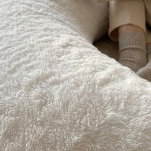 May include: A large, white, fluffy body pillow with a soft, textured surface. The pillow is crescent-shaped and appears to be made of a plush material. The image suggests a focus on comfort and relaxation.