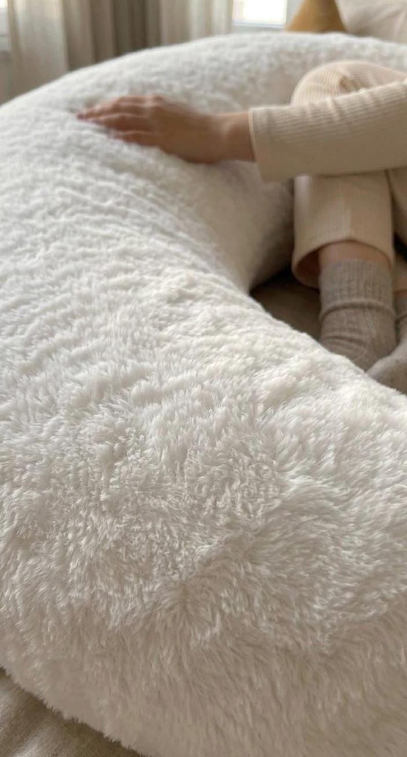 May include: A large, white, fluffy body pillow with a soft, textured surface. The pillow is crescent-shaped and appears to be made of a plush material. The image suggests a focus on comfort and relaxation.