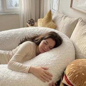 May include: A person is resting on a large, white, fluffy body pillow. A plush burger pillow and a star-shaped pillow are nearby. A mug of coffee sits on a small table. The scene is in a well-lit bedroom.