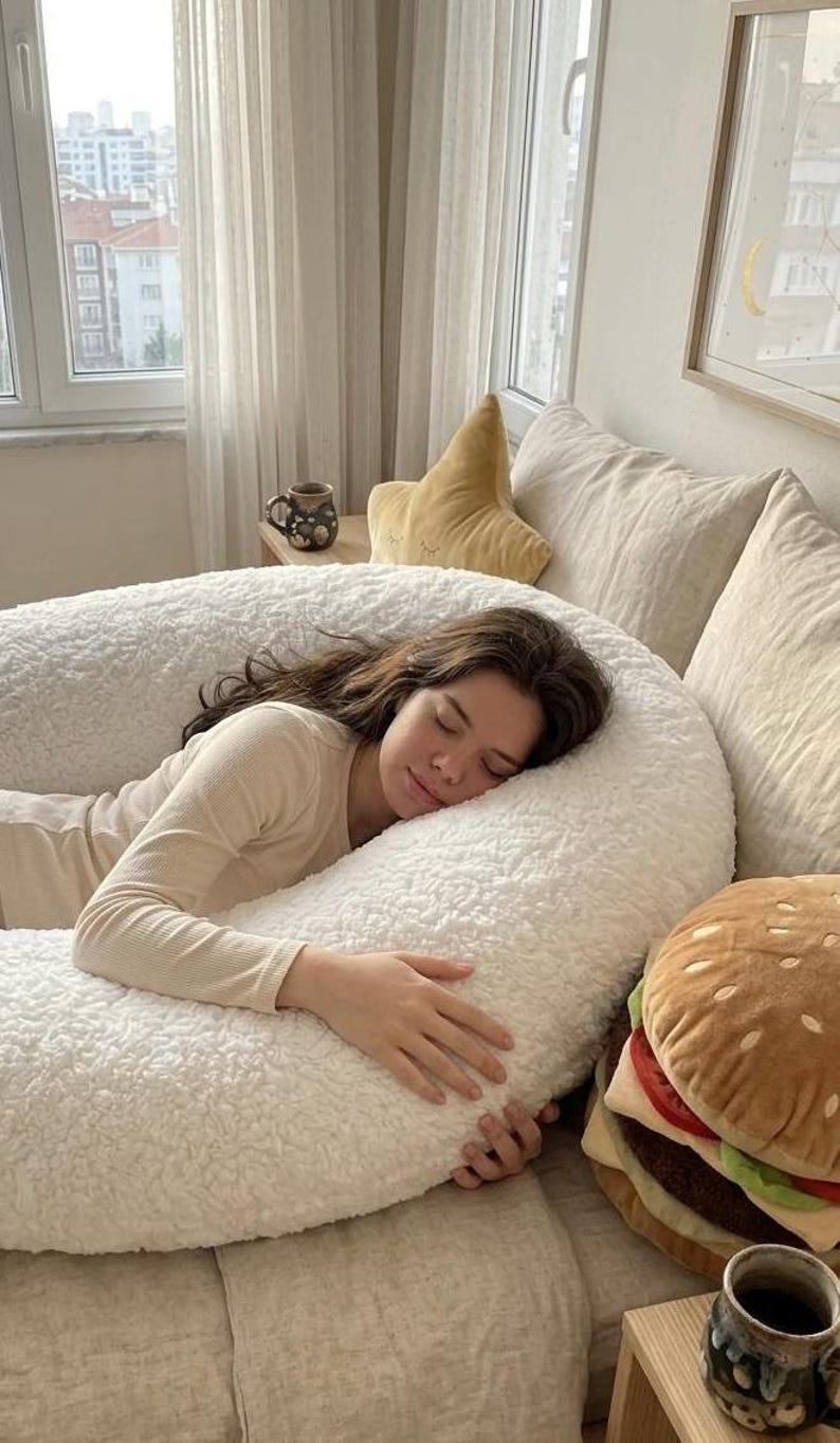 May include: A person is resting on a large, white, fluffy body pillow. A plush burger pillow and a star-shaped pillow are nearby. A mug of coffee sits on a small table. The scene is in a well-lit bedroom.