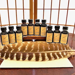 May include: A collection of eleven small glass bottles with black caps and tan labels, each featuring the text "Coven's Own Ritual Supplies" and specific names like "Attraction," "Love," and "Protection," arranged behind a large brown and white striped feather.