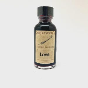 May include: A small clear glass bottle filled with dark red liquid, topped with a black screw cap. The label features the text Ceridwen's Scribe Supplies, Love, and 1 fl oz external use only, along with a small illustration of a quill pen.