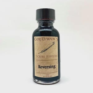 May include: A small glass bottle filled with dark liquid, featuring a black screw-top cap and a tan label. The label displays the text Ceridwen's Scribe Supplies, Reversing, and 2 fl oz external use only, along with a graphic of a quill pen.