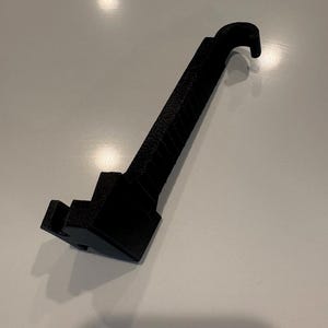 May include: A black, 3D-printed tool with a hook-shaped end and a textured handle. The tool has a stepped base and appears to be made of a matte material. The object is designed for a specific purpose, likely for lifting or manipulating objects.