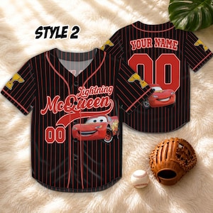Puede incluir: Camiseta de b&eacute;isbol negra con finas rayas rojas, con gr&aacute;ficos de Lightning McQueen y el n&uacute;mero 00. La camiseta tiene el texto "STYLE 2" en la parte superior izquierda. Un guante y una pelota de b&eacute;isbol est&aacute;n en primer plano.