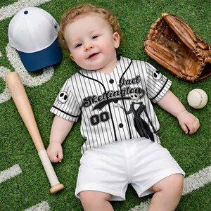 Może przedstawiać: Niemowlę w czarno-białej koszulce baseballowej w paski z nadrukiem "Jack Skellington" i "00", w połączeniu z białymi szortami. Na zdjęciu znajdują się r&oacute;wnież kij baseballowy, rękawica baseballowa, piłka baseballowa oraz biało-niebieska czapka baseballowa.