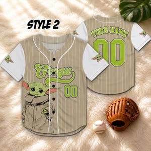 K&ouml;nnte beinhalten: Beige Baseball-Trikot mit wei&szlig;en &Auml;rmeln, mit einem Grogu-Motiv und der Nummer 00 in Gr&uuml;n. Das Trikot hat oben den Text "STYLE 2". Ein Baseballhandschuh und ein Ball sind im Bild.