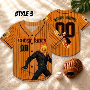 Op de afbeelding: Oranje baseballshirt met zwarte krijtstrepen, met de tekst "GHOST RIDER" en het nummer "00". Het shirt heeft een Ghost Rider-afbeelding en schedelaccenten. Een honkbalhandschoen en -bal zijn ook zichtbaar.