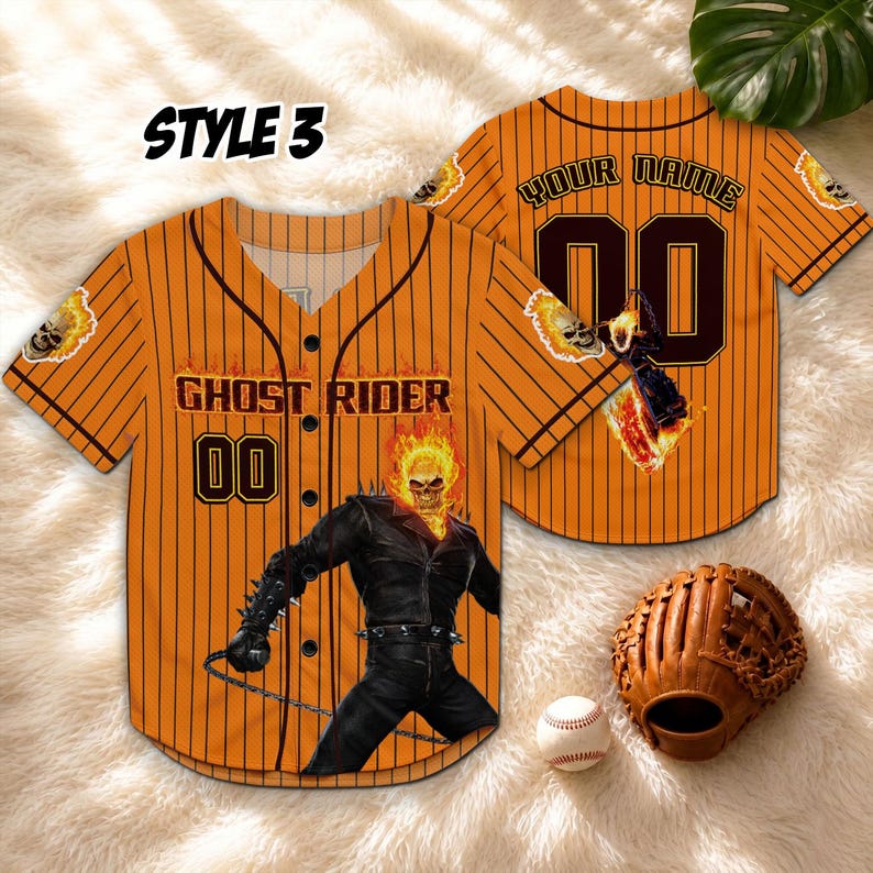 Op de afbeelding: Oranje baseballshirt met zwarte krijtstrepen, met de tekst "GHOST RIDER" en het nummer "00". Het shirt heeft een Ghost Rider-afbeelding en schedelaccenten. Een honkbalhandschoen en -bal zijn ook zichtbaar.