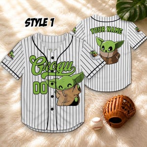 May include: Two white baseball jerseys with black pinstripes. One jersey has "Grogu" and "00" in green, with a Grogu graphic. The other jersey has "Your Name" in green, with a Grogu graphic. A brown baseball glove and a baseball are also visible.
