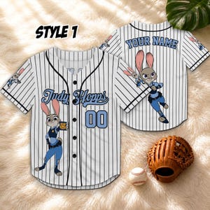 May include: Two white baseball jerseys with black pinstripes. One jersey features the text "Judy Hopps" and the number "00" in blue. The other jersey has "YOUR NAME" in blue. A baseball glove and ball are also visible.
