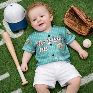 May include: A baby wearing a teal baseball jersey with the name "Rosalina" and the number "00" on the front. The jersey has white pinstripes and is paired with white shorts. A baseball bat, baseball glove, baseball, and a white and blue baseball cap are also in the image.