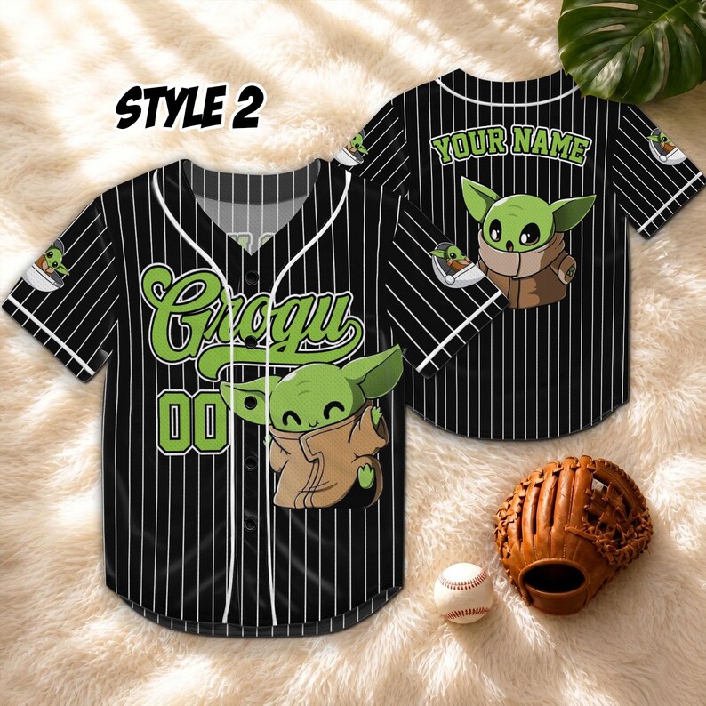 May include: Black baseball jersey with white pinstripes, featuring a Grogu graphic and the number 00. The jersey has the text "Grogu" in green script. A baseball glove and ball are next to the jersey. The jersey is labeled "Style 2".