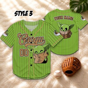 May include: Green baseball jersey with Grogu and "00" printed on the front. The jersey has vertical stripes and a brown collar. The back of the jersey has "YOUR NAME" printed above a Grogu graphic. A baseball glove and ball are next to the jersey.