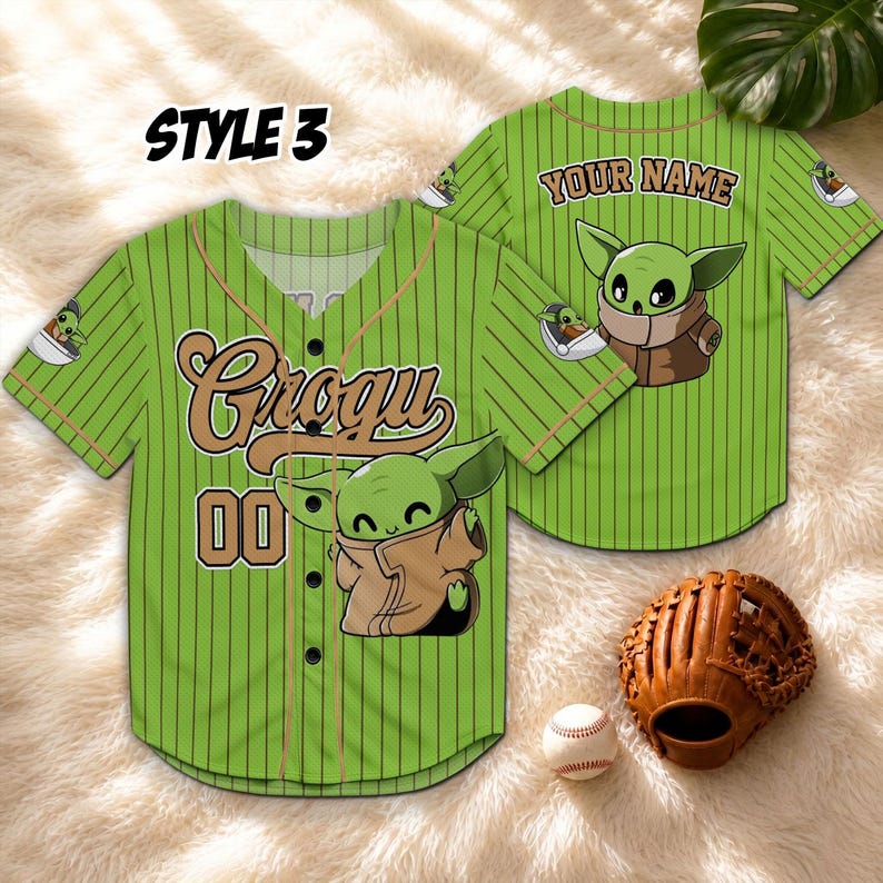 May include: Green baseball jersey with Grogu and "00" printed on the front. The jersey has vertical stripes and a brown collar. The back of the jersey has "YOUR NAME" printed above a Grogu graphic. A baseball glove and ball are next to the jersey.