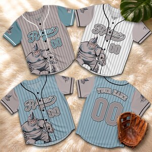 May include: Four baseball jerseys featuring a cartoon rhino design. The jerseys are in grey, white, and blue with pinstripes. Each jersey displays the word "Rhino" and the number "00". A brown baseball glove is also visible in the image.