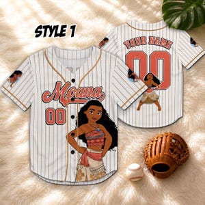 Może przedstawiać: Biała koszulka baseballowa z brązowymi prążkami, z napisem "Moana" i numerem "00" w kolorze koralowym. Koszulka ma kresk&oacute;wkową ilustrację Moany. Widoczna jest druga koszulka, a także rękawica i piłka do baseballu.