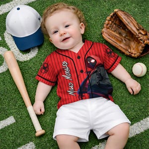 May include: A baby wearing a red and black pinstripe baseball jersey with "Miles Morales" printed on the front. The jersey features a graphic of a superhero. The baby is also wearing white shorts. A baseball bat, baseball glove, baseball, and a white and blue baseball cap are nearby.