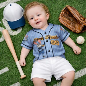 May include: A baby wearing a light blue baseball jersey with the text "Judy Hopps" and the number "00". The jersey has a cartoon image of a rabbit. The baby is also wearing white shorts. A baseball bat, baseball glove, baseball, and a white and blue baseball cap are also in the image.