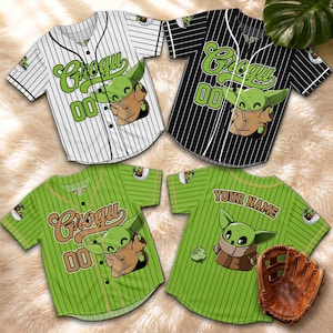 May include: Four baseball jerseys featuring Grogu graphics. The jerseys are white with black pinstripes, black with white pinstripes, and green with green pinstripes. One green jersey has a baseball glove. The jerseys have the number 00.