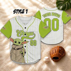K&ouml;nnte beinhalten: Wei&szlig;es und gr&uuml;nes Baseball-Trikot mit dem Text "Grogu" und der Nummer "00". Das Trikot zeigt ein Cartoon-Bild von Grogu. Die &Auml;rmel sind gr&uuml;n mit einem Grogu-Bild. Auf der R&uuml;ckseite des Trikots steht der Text "YOUR NAME" und die Nummer "00".