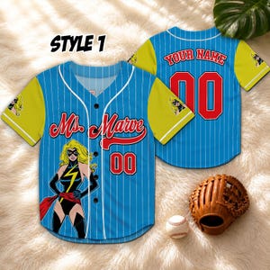 May include: Blue baseball jersey with white pinstripes, yellow sleeves, and a Ms. Marvel graphic. The jersey features the text "Ms. Marvel" and the number "00" in red. A baseball glove and ball are also visible.