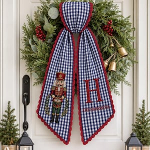 May include: A blue and white gingham fabric door hanger featuring a red lace trim. One side displays an embroidered nutcracker soldier playing a drum, while the other side features a large red letter H and the text THE HARRISONS embroidered below.