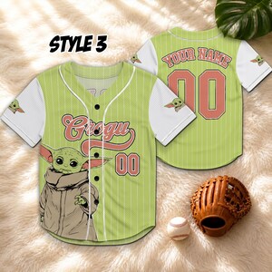 K&ouml;nnte beinhalten: Gr&uuml;nes und wei&szlig;es Baseball-Trikot mit dem Text "Grogu" und der Nummer "00" auf der Vorderseite. Das Trikot zeigt eine Illustration von Grogu. Auf der R&uuml;ckseite des Trikots steht der Text "YOUR NAME" und die Nummer "00". Ein Baseballhandschuh und ein Ball sind im Bild.