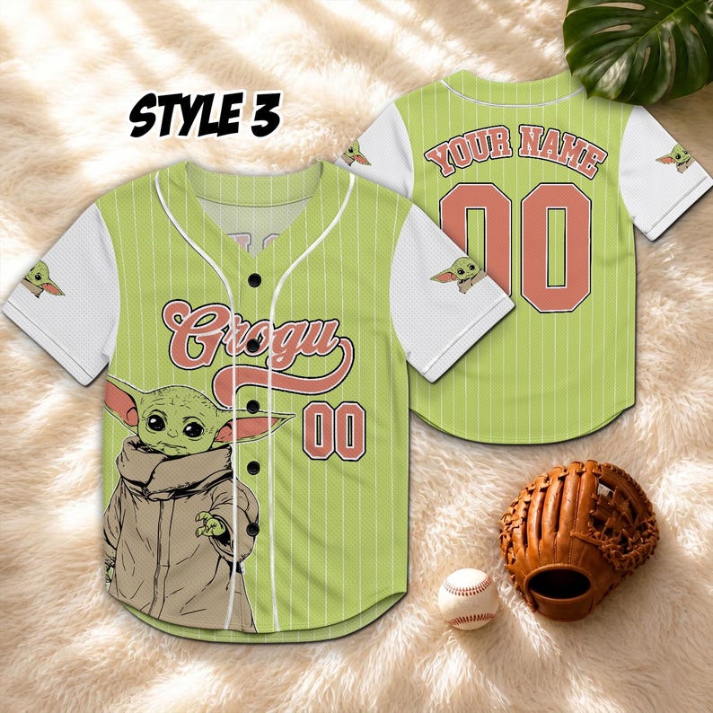K&ouml;nnte beinhalten: Gr&uuml;nes und wei&szlig;es Baseball-Trikot mit dem Text "Grogu" und der Nummer "00" auf der Vorderseite. Das Trikot zeigt eine Illustration von Grogu. Auf der R&uuml;ckseite des Trikots steht der Text "YOUR NAME" und die Nummer "00". Ein Baseballhandschuh und ein Ball sind im Bild.