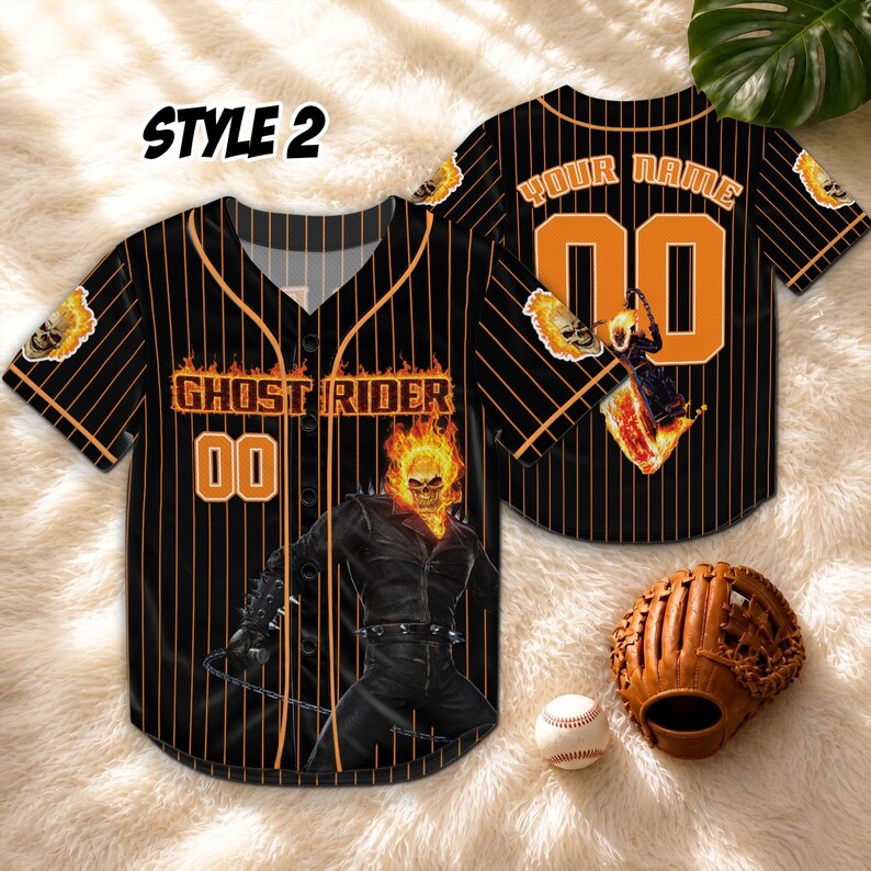 Op de afbeelding: Zwart honkbalshirt met oranje krijtstrepen en de woorden "GHOST RIDER" en "00" in oranje. Het shirt heeft een afbeelding van Ghost Rider en een vlammende schedel. Een honkbal, handschoen en de woorden "STYLE 2" zijn ook zichtbaar.