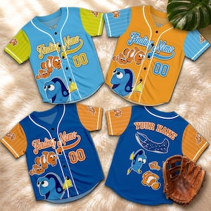 Op de afbeelding: Vier honkbalshirts in blauw, oranje en geel met "Finding Nemo" graphics. Elk shirt heeft het nummer "00" en stripfiguren. Eén shirt heeft "YOUR NAME" op de achterkant. Een bruine honkbalhandschoen is ook zichtbaar.