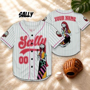 Może przedstawiać: Biała koszulka baseballowa z pionowymi prążkami, z napisem "SALLY" w czerwonym skrypcie i numerem "00". Koszulka ma kresk&oacute;wkową ilustrację Sally z The Nightmare Before Christmas. Widoczna jest r&oacute;wnież rękawica i piłka do baseballu.