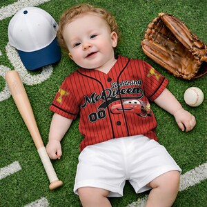Puede incluir: Un beb&eacute; con una camiseta de b&eacute;isbol roja con "Lightning McQueen" y "00" impresos, combinada con pantalones cortos blancos. Un bate de b&eacute;isbol, un guante, una pelota y una gorra de b&eacute;isbol blanca y azul tambi&eacute;n est&aacute;n presentes en un campo verde.