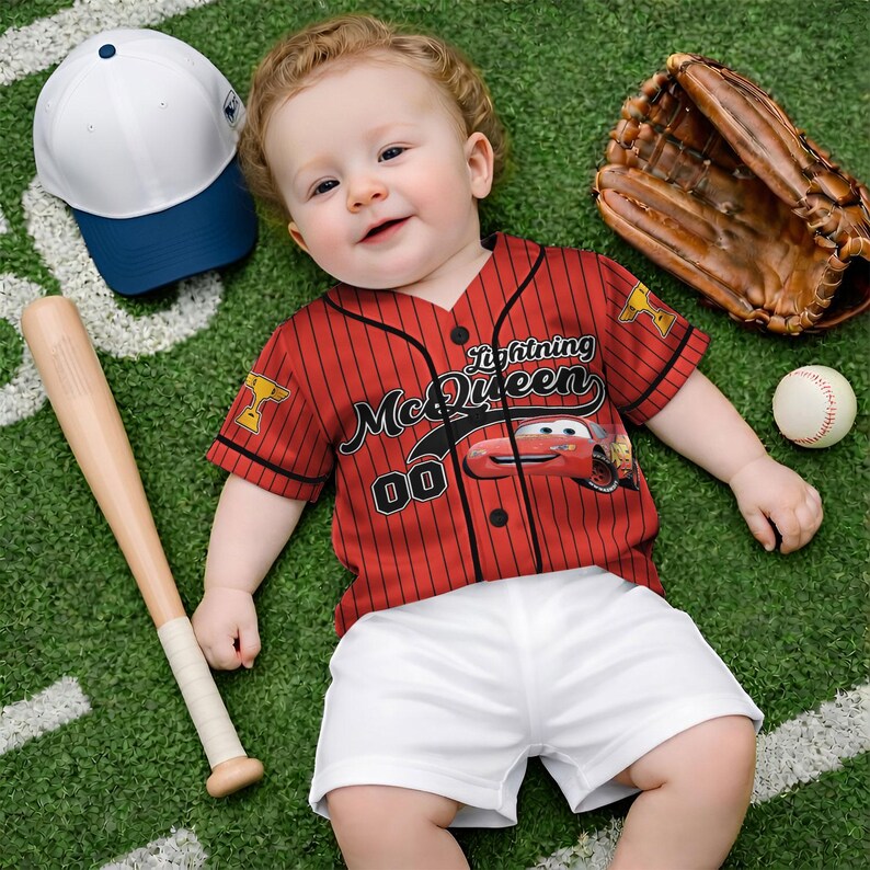 Puede incluir: Un beb&eacute; con una camiseta de b&eacute;isbol roja con "Lightning McQueen" y "00" impresos, combinada con pantalones cortos blancos. Un bate de b&eacute;isbol, un guante, una pelota y una gorra de b&eacute;isbol blanca y azul tambi&eacute;n est&aacute;n presentes en un campo verde.