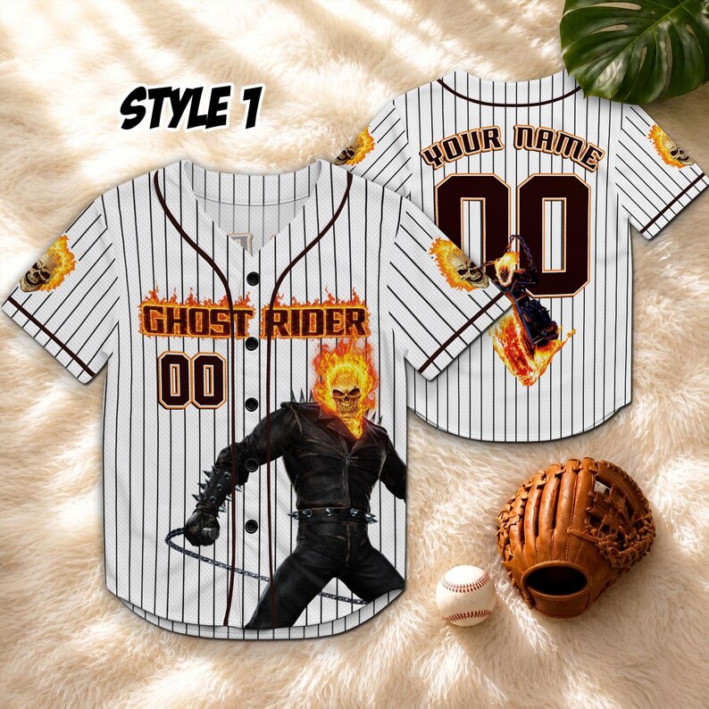 Op de afbeelding: Wit honkbalshirt met zwarte krijtstrepen, met een Ghost Rider-afbeelding. Het shirt heeft de tekst "GHOST RIDER" en het nummer "00" in een vurige lettertype. Een honkbalhandschoen en -bal zijn ook zichtbaar.