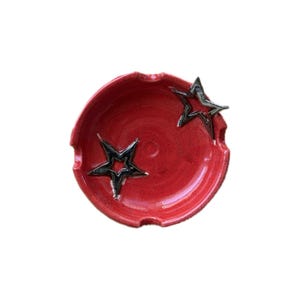 May include: Red ceramic ashtray with two star-shaped accents. The ashtray has a circular shape with three notches and a glossy finish. The stars are black with silver outlines.